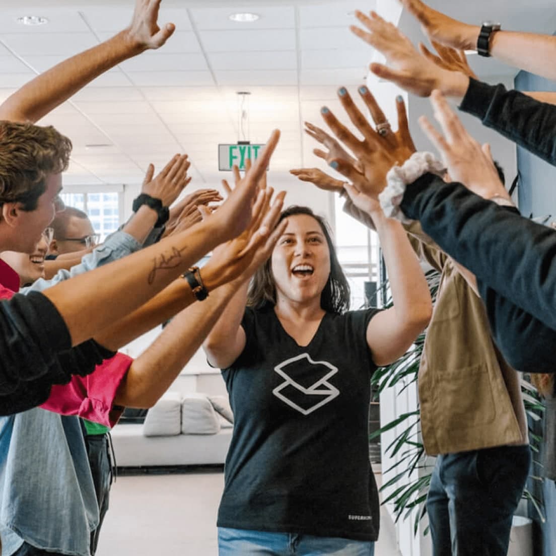 Enthusiastic group celebrating with raised hands in an office space; woman in black shirt with logo at centre.