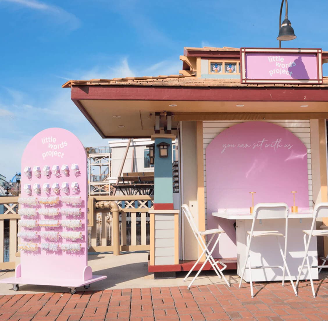 Pink-themed "Little Words Project" retail kiosk with bracelet display and outdoor seating area under blue sky.
