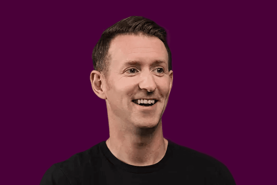 A smiling man in a black t-shirt clasps his hands together against a deep purple background.